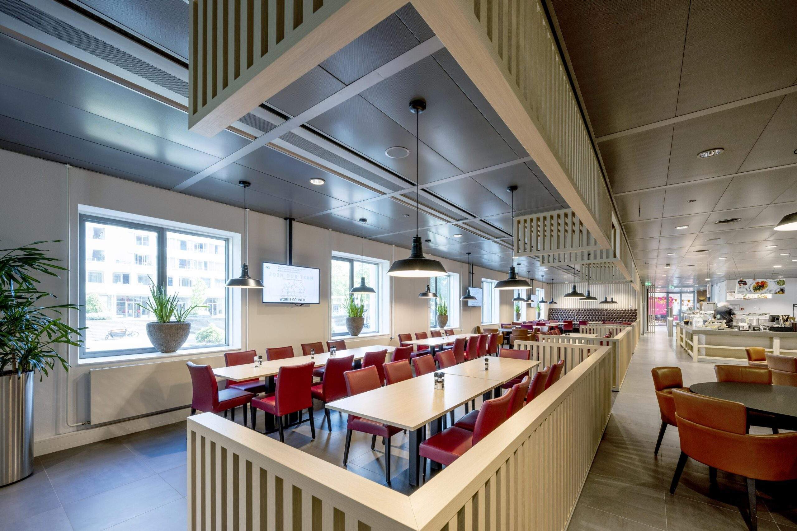 Moderne en ruim opgezette bedrijfskantine aan het Koningin Wilhelminaplein met roodbruine stoelen, houten details en grote ramen.